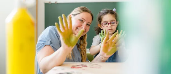 Eine Erzieherin zeigt ihre bunten Hände beim Malen mit einem Kind
